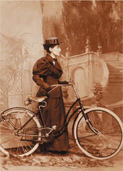 Annie standing with her bicycle in a full Victorian-style sleeved dress and hat